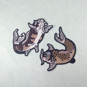 以下が含まれることがあります： 2つの鯉の刺繍パッチ。1匹の魚は黒、茶色、白で、詳細なデザインです。もう1匹は黒と茶色で、詳細な鱗模様です。どちらのパッチも黒い縁取りがあり、薄緑色の背景にあります。