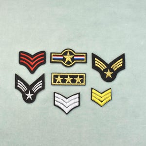 May include: A collection of seven embroidered military-style patches in various shapes and colours. The set includes chevron designs in red, white, and yellow, a rectangular patch with three gold stars, and a circular patch featuring a central star.