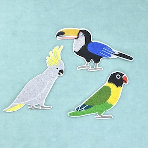 Peut inclure: Trois écussons brodés représentant des oiseaux : un cacatoès gris à crête jaune, un toucan au grand bec jaune et orange avec des ailes bleues, et un inséparable vert à face noire et bec orange, tous dotés de coutures détaillées.