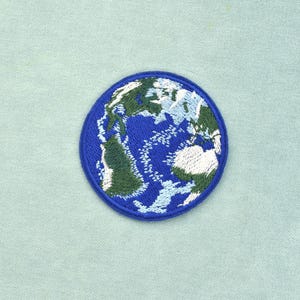 Peut inclure: Un écusson brodé circulaire représentant la Terre. Le motif présente des océans bleu foncé, des masses terrestres vert foncé et des formations nuageuses blanches, le tout entouré d'une bordure cousue bleu uni sur un tissu clair.