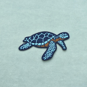 May include: An embroidered patch in the shape of a sea turtle. The turtle features a dark navy blue shell with light blue geometric patterns, a light blue head and flippers with dark navy outlines, and a brown underside detail on the body.