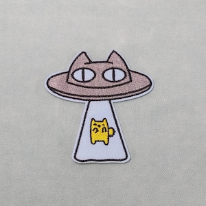 Peut inclure: Patch brodé représentant un OVNI en forme de chat brun avec un chat jaune à l'intérieur. L'OVNI a de grands yeux noirs.