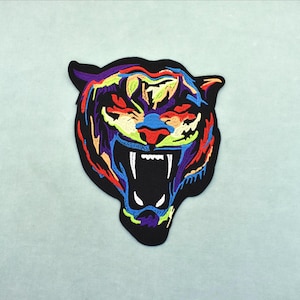 May include: An embroidered tiger head patch with a black outline and a vibrant, multi-coloured design. The tiger's face is filled with shades of red, blue, yellow, and purple. This patch is designed to be applied to clothing or accessories.