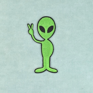 May include: An embroidered patch featuring a bright green alien character with a large oval head and black almond-shaped eyes. The alien is depicted in a standing position, raising one arm to make a peace sign gesture with its hand.