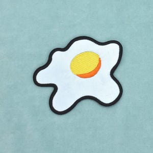 May include: An embroidered patch shaped like a fried egg with a white egg white section and a central yellow and orange yolk. The entire shape is outlined with a thick black border, creating a distinct contrast against the light blue background.