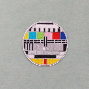 Könnte beinhalten: Ein runder, weißer gestickter Aufnäher mit einem schwarz-weißen Fernseh-Testmuster. Das Muster enthält farbige Balken in Rot, Blau, Grün, Gelb, Rosa und Cyan.