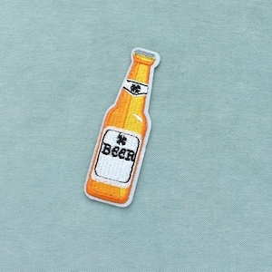 May include: Embroidered patch of a beer bottle in orange and yellow, with a white label reading "BEER" in black. The patch has a white border and is set against a light blue background.