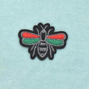 以下が含まれることがあります： 黒い体に白いステッチのディテールが施された、刺繍入りのハチのワッペン。羽は2つの水平なセクションに分かれており、上半分は鮮やかな赤、下半分は緑色で、全体がしっかりとした黒い縁取りで囲まれています。