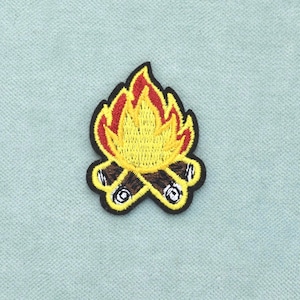 Op de afbeelding: Een geborduurde patch met een kampvuurontwerp met bruine houtblokken aan de basis en felgele en rode vlammen erboven. De gehele vorm is omlijnd met een dikke zwarte rand, wat zorgt voor een duidelijke en heldere grafische weergave.