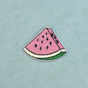 Peut inclure: Un écusson brodé en forme de tranche de pastèque. Le fruit présente un intérieur rose avec des pépins noirs brodés, une fine écorce blanche et un bord extérieur vert, le tout souligné par une bordure sombre sur un fond bleu clair.