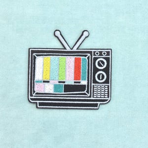 Puede incluir: Un parche bordado en blanco y negro con forma de televisor antiguo. La pantalla muestra un patrón de prueba colorido con rayas verticales en amarillo, verde, rosa y rojo, junto con dos antenas que se extienden desde la parte superior.