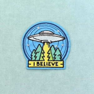 Peut inclure: Un écusson brodé circulaire représentant un OVNI argenté planant au-dessus de sapins verts. Un faisceau lumineux jaune descend du vaisseau. Le texte I BELIEVE est brodé en noir sur une bannière rectangulaire jaune au bas de l'écusson.
