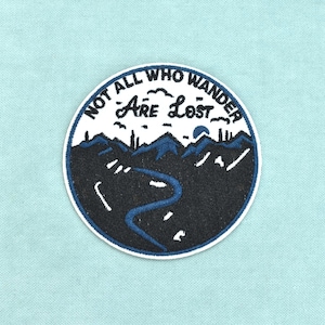 May include: A circular embroidered patch featuring a dark blue mountain landscape with a winding river and a sun. The text "NOT ALL WHO WANDER ARE LOST" is stitched in black along the top edge of the white sky area within the circular border.