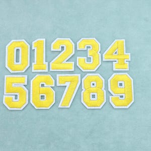 Può includere: Un set di dieci toppe termoadesive individuali raffiguranti i numeri da zero a nove. Ogni numero è realizzato in tessuto giallo brillante con un bordo ricamato bianco spesso, progettato in un classico carattere in stile universitario.