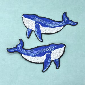 Puede incluir: Dos parches bordados de ballena con un cuerpo superior azul vibrante y una parte inferior blanca. Cada ballena está delineada con un borde negro grueso, mostrando costuras detalladas que definen las aletas, la cola y el ojo con precisión.