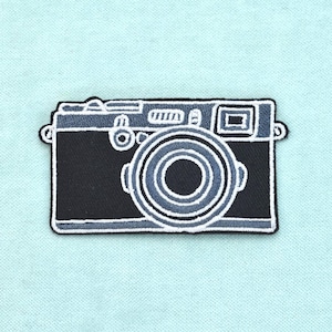 May include: An embroidered patch featuring a black and grey camera design with white outlines. The camera is depicted from a front-facing perspective, showcasing a prominent circular lens, a rectangular viewfinder, and various buttons.