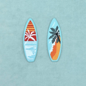 Op de afbeelding: Twee geborduurde surfplank-patches met lichtblauwe randen. De linker patch toont het silhouet van een witte palmboom tegen een rode en oranje zonsondergang boven een blauwe oceaan. De rechter patch toont een donkergroene palmboom boven een zandstrand.