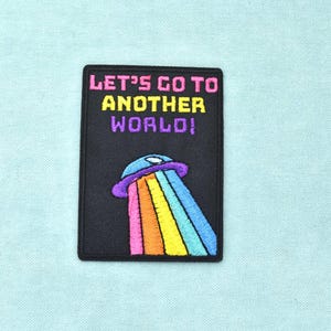 Peut inclure: Un écusson rectangulaire brodé noir avec le texte LET'S GO TO ANOTHER WORLD! en rose, jaune et violet. Sous le texte se trouve un OVNI violet et bleu émettant un large faisceau lumineux arc-en-ciel aux rayures verticales colorées.