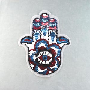 Peut inclure: Un écusson en forme de main de Hamsa orné de sequins aux tons rouge profond, bleu roi et blanc. L'applique présente un motif floral central et des détails complexes, le tout entouré d'une bordure en tissu blanc pour une fixation facile.