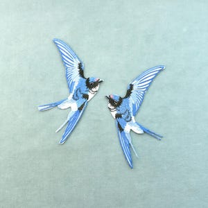 May include: Two embroidered patches of blue swallows with outstretched wings. The birds have white and black details on their wings and heads. The patches are on a light blue background.