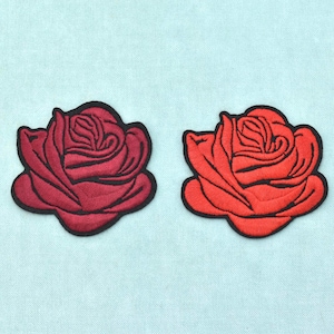 Könnte beinhalten: Zwei gestickte Rosenaufnäher mit schwarzem Rand. Ein Aufnäher ist in tiefem Burgunderrot gehalten, der andere in einem leuchtenden Rot. Beide weisen detaillierte Stickereien auf, die die geschichteten Blütenblätter einer Rose darstellen.