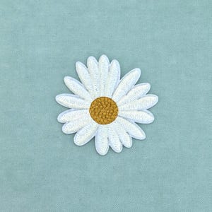 May include: A daisy flower patch featuring white embroidered petals arranged in a circular pattern around a textured, golden-yellow center. The embroidery is dense and uniform, creating a clear floral shape with defined edges and a raised surface.