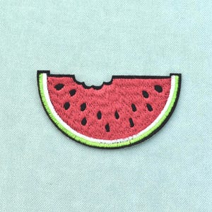 May include: An embroidered patch shaped like a slice of watermelon with a bite taken out of the top. The patch features a red center with black seeds, a white rind, and a bright green outer edge, all outlined in a thick black thread border.