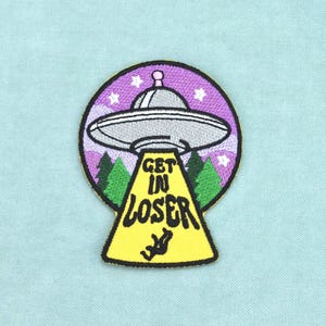 Peut inclure: Un écusson brodé circulaire représentant un OVNI gris survolant des pins verts sous un ciel violet étoilé. Un rayon tracteur jaune s'étend vers le bas avec le texte noir GET IN LOSER et une petite silhouette noire d'une personne.