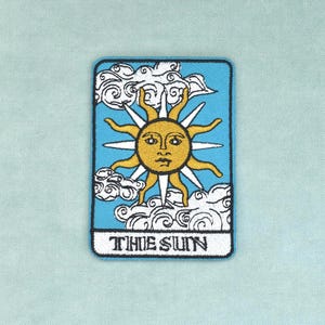 May include: A rectangular embroidered patch featuring a yellow sun with a face, surrounded by white clouds against a light blue background. The sun has alternating straight and wavy rays. The text THE SUN is embroidered in black at the bottom.