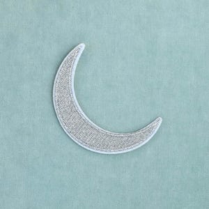 Pode incluir: Um emblema em forma de meia-lua com uma superfície bordada em prata metálica e uma borda costurada em branco. O design tem a forma de uma lua fina e curva, exibindo um padrão de fio texturizado em toda a forma da meia-lua prateada.