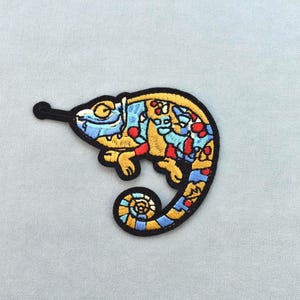 May include: Embroidered patch of a chameleon with a black border and yellow outline. The chameleon's body is filled with blue, red, and yellow patterns. The patch is approximately 7.6 cm long.
