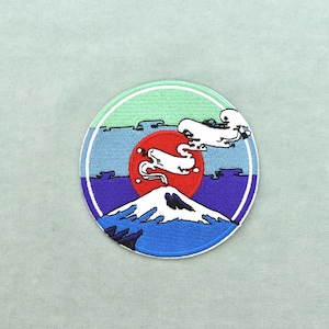 May include: A circular embroidered patch featuring a snow-capped mountain peak in white and blue at the bottom. Above the mountain, a large red sun sits against a purple and blue sky, with white stylized clouds and a light green upper border.