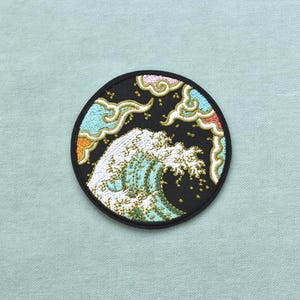 Pode incluir: Um emblema bordado circular apresentando uma onda oceânica branca estilizada sobre um fundo preto. O design inclui nuvens azul-claras, amarelas e rosas com contornos dourados ao redor da onda, tudo envolvido por uma borda preta.
