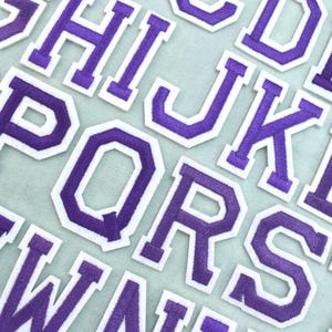 May include: A collection of iron-on alphabet patches featuring purple letters with a white border in a classic varsity block font. The letters are arranged in rows, showcasing the full English alphabet from A to Z in a consistent style.