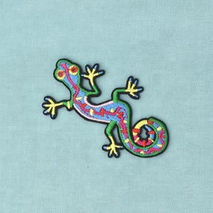Pode incluir: Um emblema bordado apresentando um lagarto estilizado com contorno preto. O lagarto tem um corpo azul-claro com membros verde-brilhantes, dedos amarelos e um padrão vibrante de detalhes em vermelho, rosa e amarelo ao longo das costas.