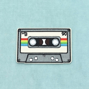 Op de afbeelding: Een geborduurde patch in de vorm van een rechthoekige cassetteband. Het ontwerp heeft een grijs frame, een wit label in het midden en een horizontale regenboogstreep. De tekst SIDE B en het getal 90 zijn in het zwart in de hoeken geborduurd.