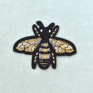 Peut inclure: Un écusson brodé en forme d'abeille avec une tête et un corps noirs, agrémenté de fils dorés et bleus sur les ailes et l'abdomen. Le motif de l'insecte est souligné par un contour en fil noir, créant une forme nette sur le tissu clair.