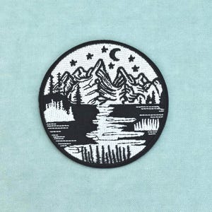 以下が含まれることがあります： 黒と白の風景デザインが施された円形の刺繍ワッペン。三日月と星の下に山脈が描かれ、手前には松林の中を流れる川が表現されており、全体が黒い縁取りで囲まれています。