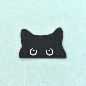 Pode incluir: Um emblema bordado preto em forma de cabeça de gato com orelhas pontiagudas. O emblema apresenta dois olhos circulares brancos com pequenas pupilas brancas, criando uma expressão de espreita. O design é plano com uma borda costurada.