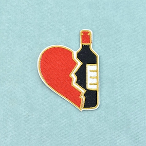 Peut inclure: Un écusson brodé représentant un cœur rouge divisé en deux, dont la partie droite est remplacée par une bouteille de vin noire avec un bouchon rouge et une étiquette blanche. Le motif est entièrement bordé d'un fil doré, formant un emblème.
