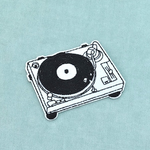 May include: A black and white embroidered patch featuring a detailed illustration of a DJ turntable. The design includes a circular record platter, a tonearm, and various control knobs and sliders, all rendered in high-contrast black thread.