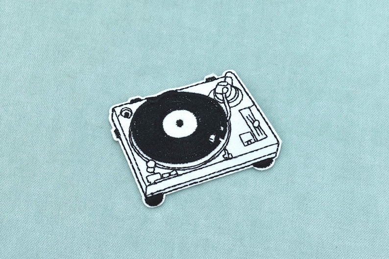 May include: A black and white embroidered patch featuring a detailed illustration of a DJ turntable. The design includes a circular record platter, a tonearm, and various control knobs and sliders, all rendered in high-contrast black thread.