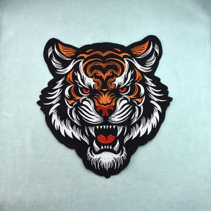May include: Embroidered tiger head patch with a black background, orange and white details, and red eyes. The tiger's mouth is open, revealing sharp white teeth and a red tongue. The patch is likely for clothing or accessories.