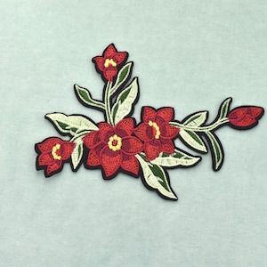 Peut inclure: Un écusson en tissu brodé représentant un groupe de fleurs rouges aux centres jaunes avec des feuilles vertes. Le motif comprend trois fleurs épanouies, deux petits boutons et plusieurs feuilles, le tout souligné par une bordure noire.