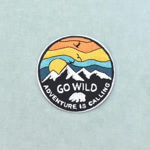 Op de afbeelding: Een ronde geborduurde patch met een berglandschap, een gele zon, blauwe lucht en oranje wolken. Onder de witte bergtoppen bevindt zich een zwart gedeelte met de witte tekst GO WILD en ADVENTURE IS CALLING en een icoon van een beer.