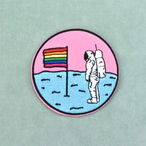 Pode incluir: Um emblema bordado circular apresentando um astronauta branco em pé sobre uma superfície lunar azul-clara. Um mastro com uma bandeira de listras horizontais de arco-íris fica ao lado do astronauta contra um céu rosa com borda preta.