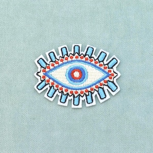 Peut inclure: Un écusson brodé en forme d'œil avec une pupille rouge centrale, un iris bleu clair et une sclérotique blanche. Le motif présente une bordure décorative composée de rayons rectangulaires bleu clair et de petits points rouges sur fond blanc.