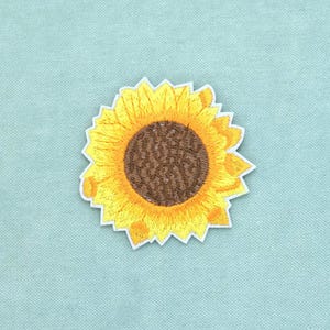 May include: A sunflower embroidered patch featuring a dark brown centre with textured stitching and bright yellow petals radiating outwards. The patch has a white die-cut border that follows the jagged, irregular shape of the flower petals.