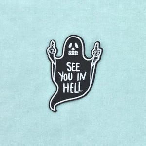 Pode incluir: Um emblema bordado preto em forma de fantasma com um rosto de caveira. O fantasma tem dois braços levantados com os dedos médios estendidos. O texto branco no centro do fantasma diz, SEE YOU IN HELL, em uma fonte negrito e maiúscula.