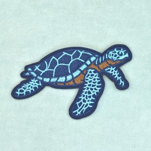 以下が含まれることがあります： ウミガメの形をした刺繍ワッペン。カメは、水色の輪郭を持つ濃い青色の甲羅、濃い青色のディテールが施された水色の頭とヒレ、そしてカメの体の茶色の腹部が特徴です。